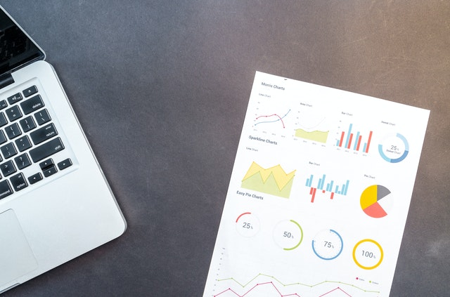 Corner of a macbook and a sheet of paper with line graphs and pie charts on a grey table